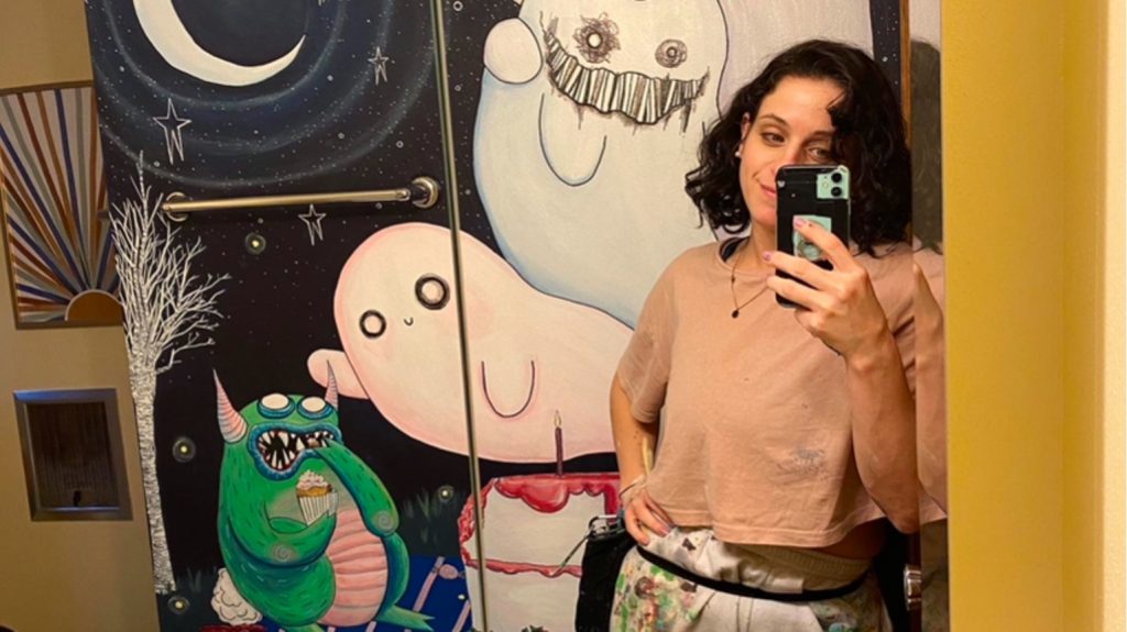 A photograph of Keighley Stadler standing in front of one of her paintings of adorable monsters.